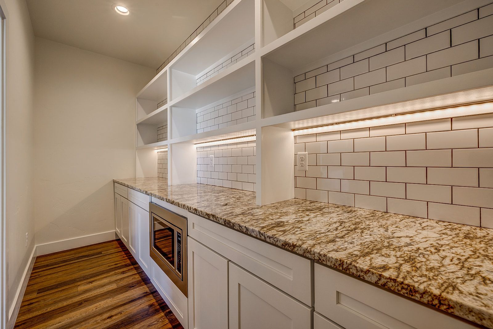 White pantry with granite countertop, built-in microwave, wood floor, and tiled backsplash.