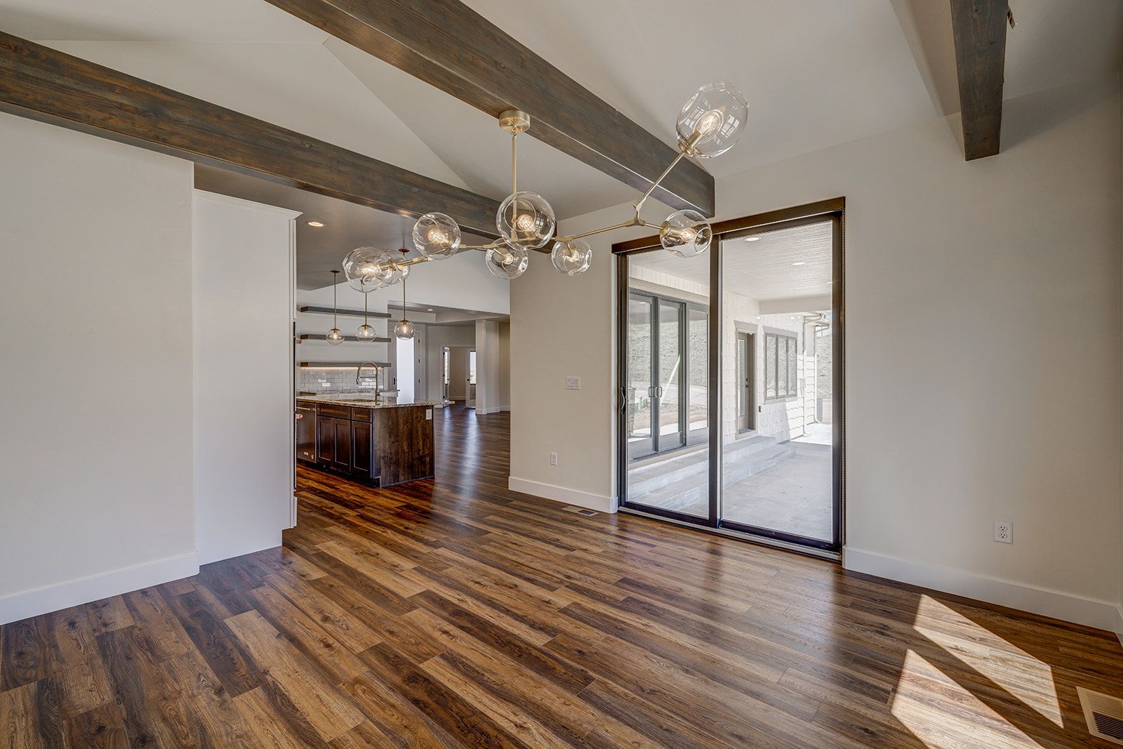 Dining room with wooden floor, exposed beams, and glass sliding doors. Kitchen visible in the background.