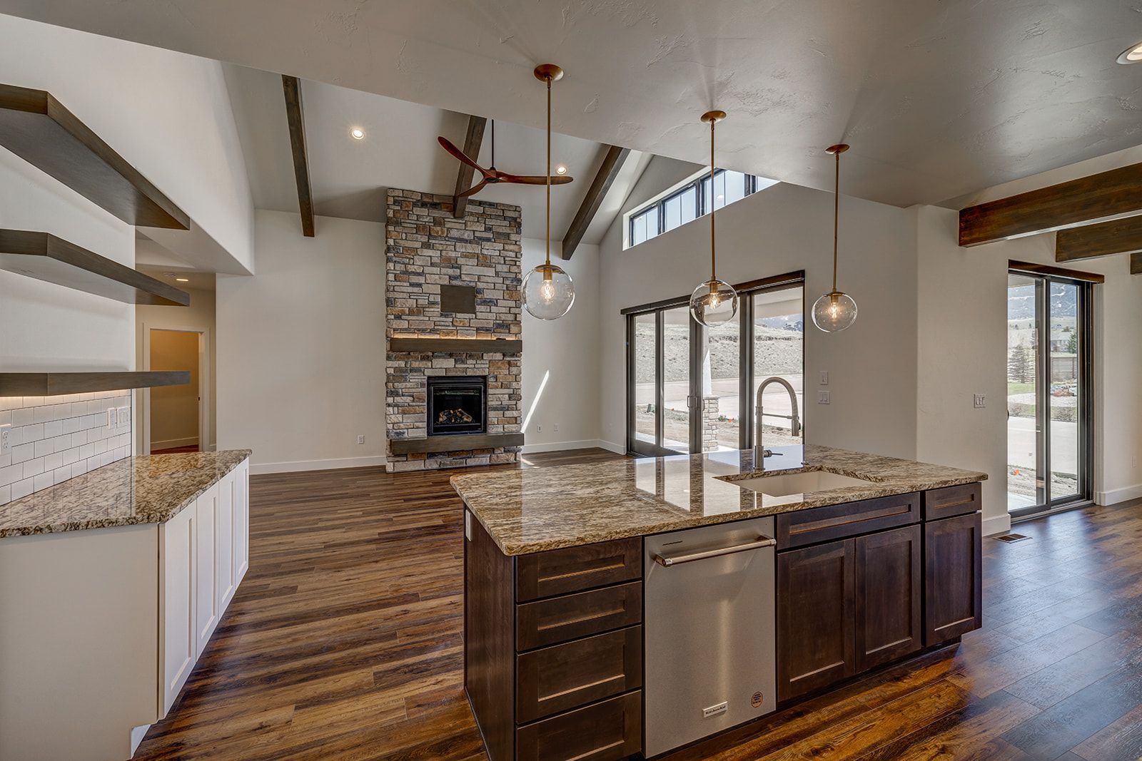 Open-concept kitchen and living area with a stone fireplace, dark wood cabinets, and large windows.