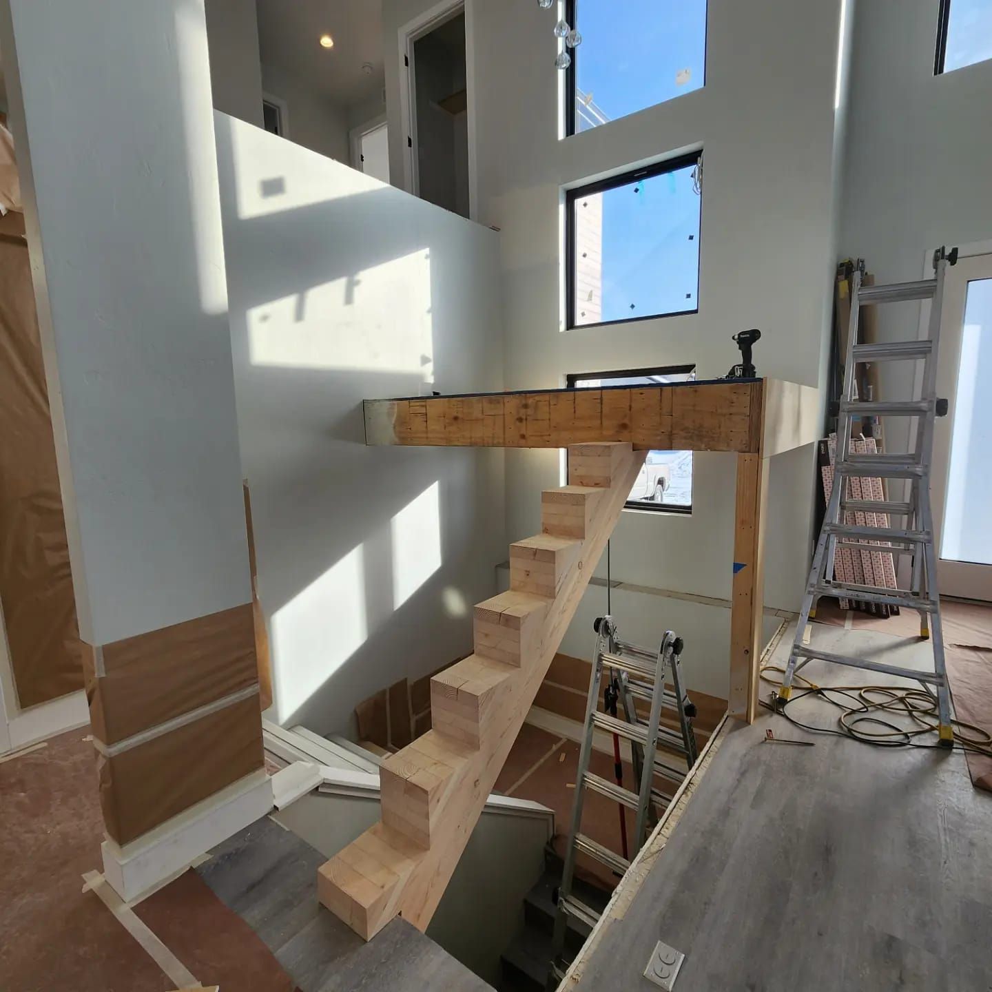 Interior of a house under construction; wooden stairs lead to a platform; tall windows, ladder, and tools present.