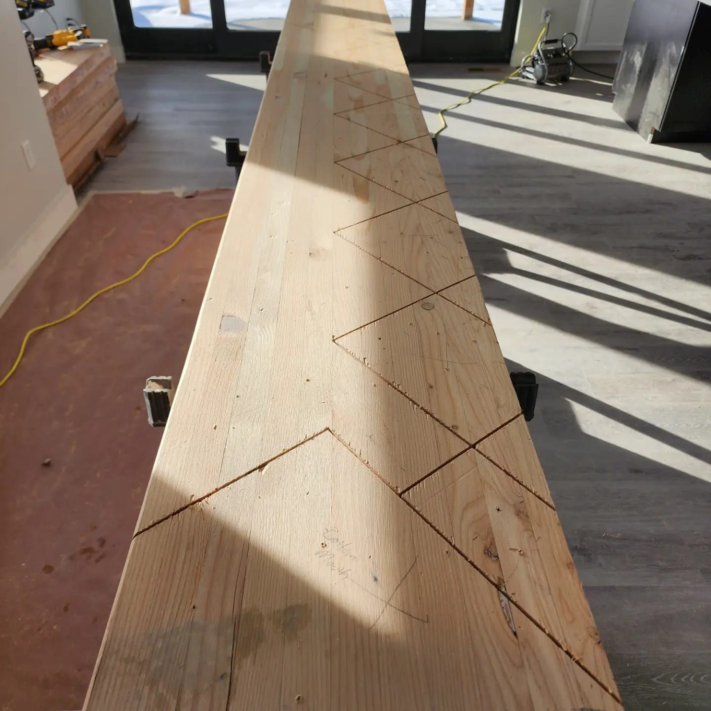 A long wooden board with zig-zag cuts, likely for stairs, indoors on sawhorses.
