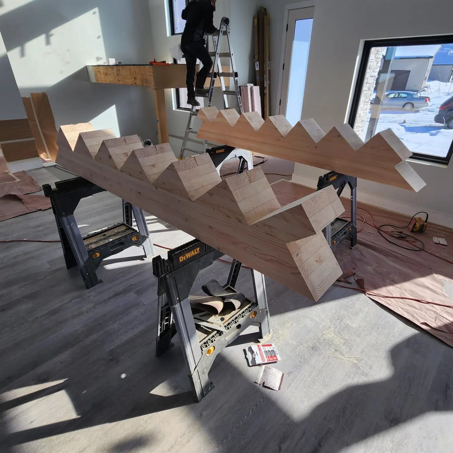 Two wooden stair stringers on sawhorses in a room under construction; a person on a ladder in the background.
