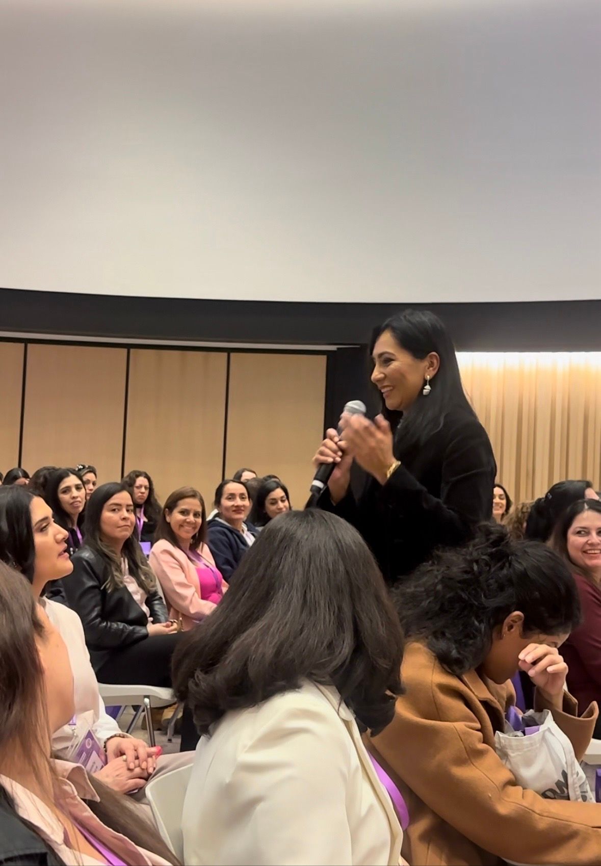 Woman speaking into a microphone, addressing a seated audience in a large room.