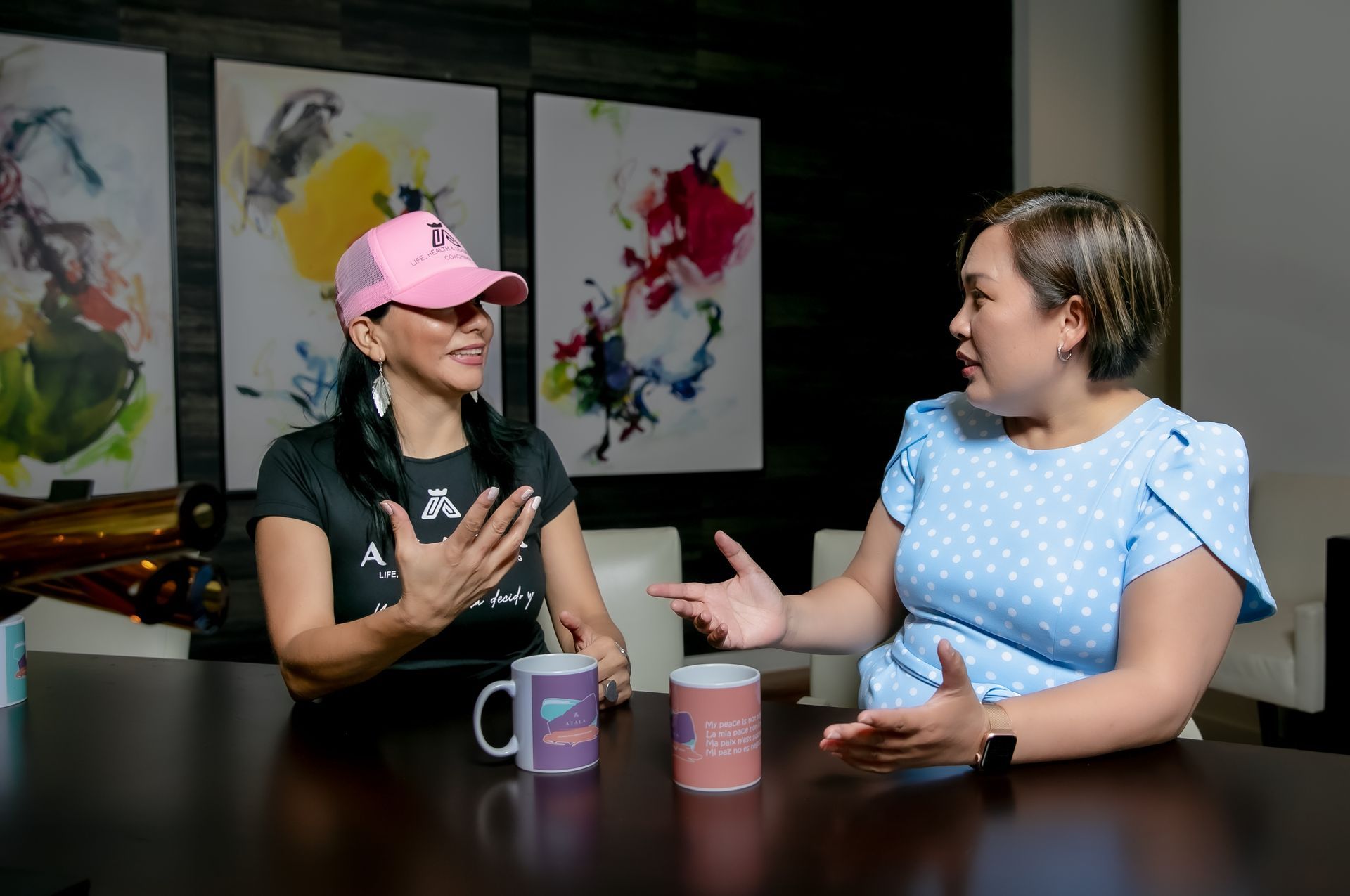 Two people in a modern room with abstract art talk while sitting at a dark table with mugs.