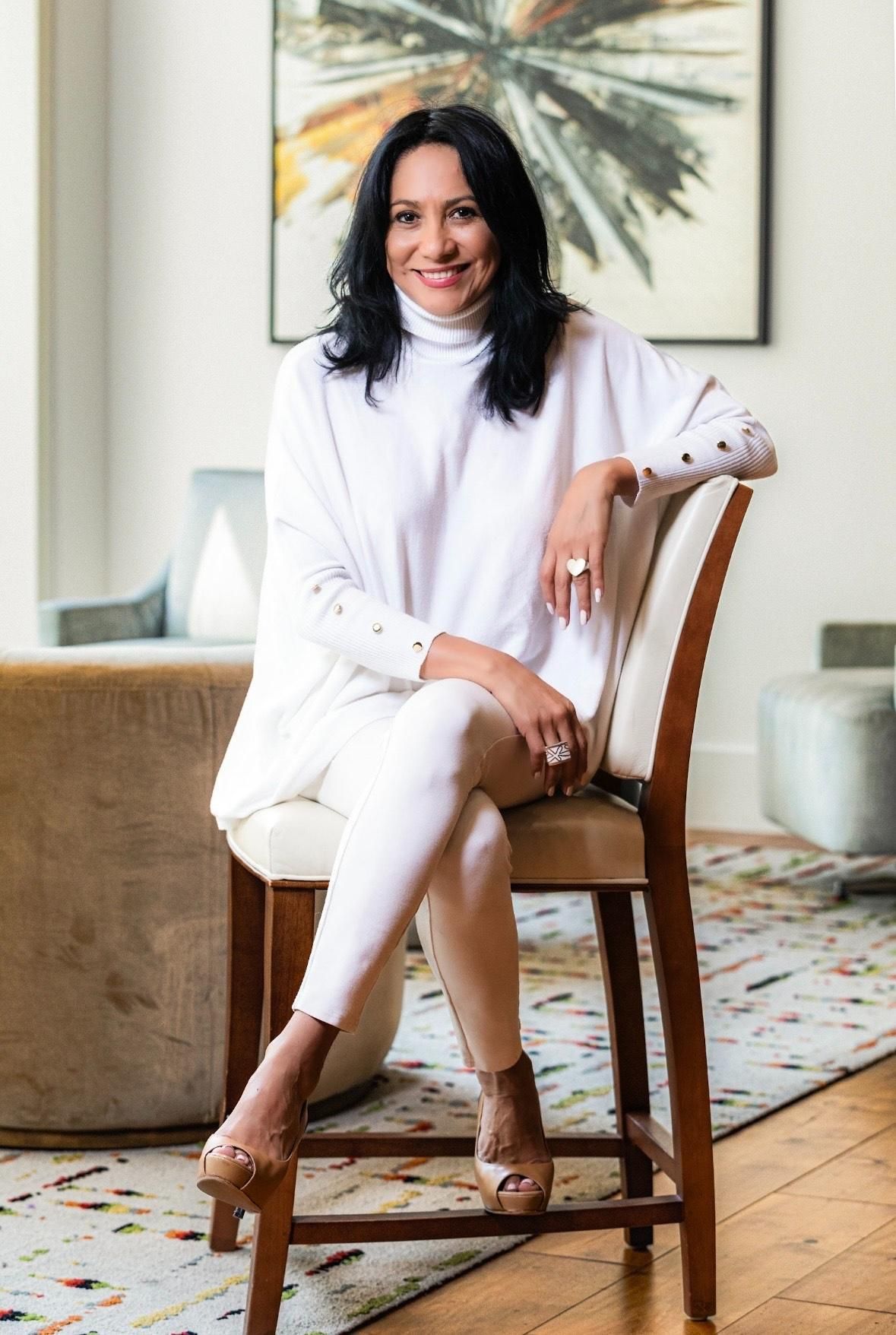 A person sitting on a chair in a bright, modern room, wearing a white turtleneck and pants, smiling at the camera.