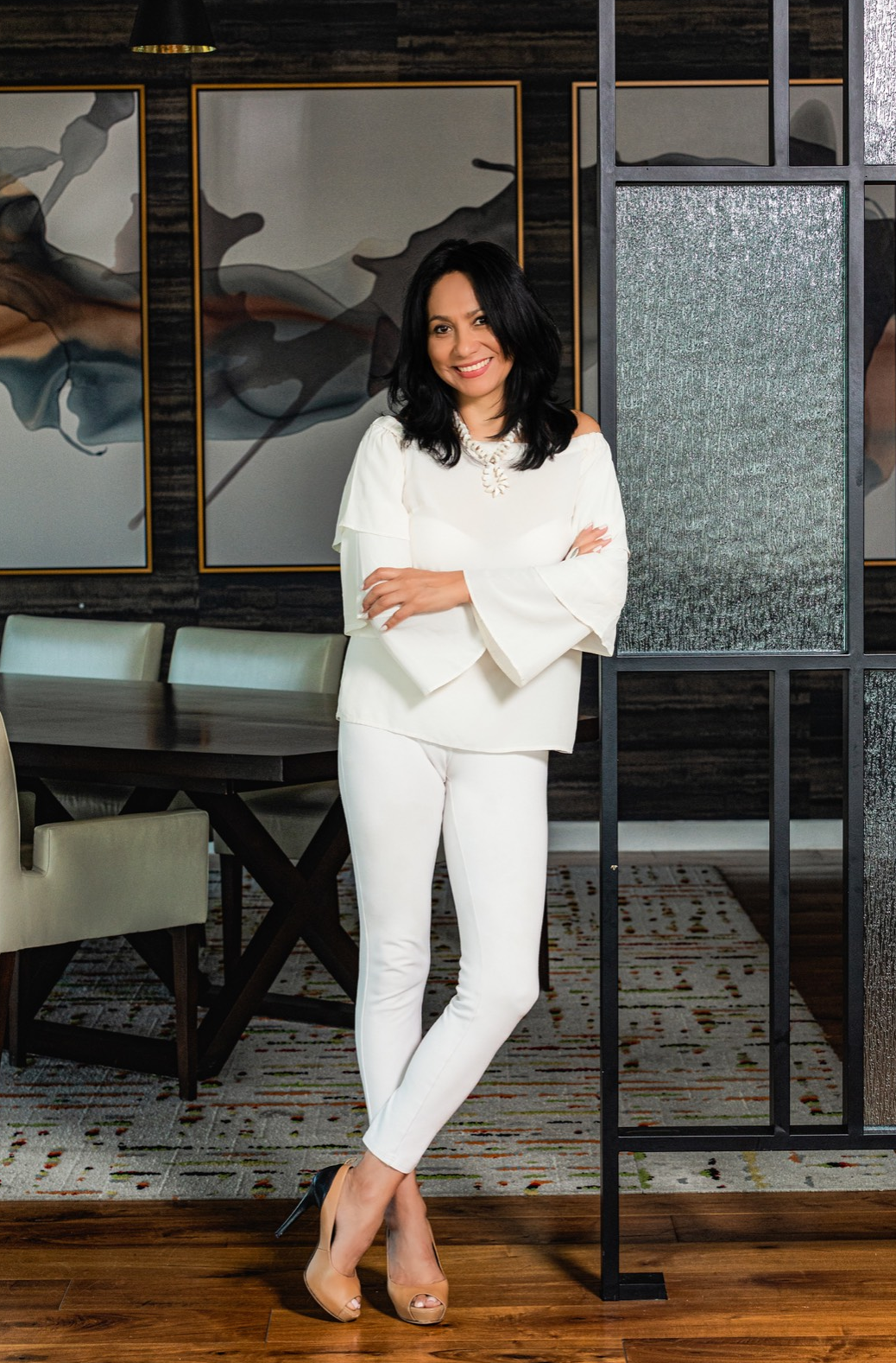 Woman in white outfit, arms crossed, poses near a room divider, dining table background.