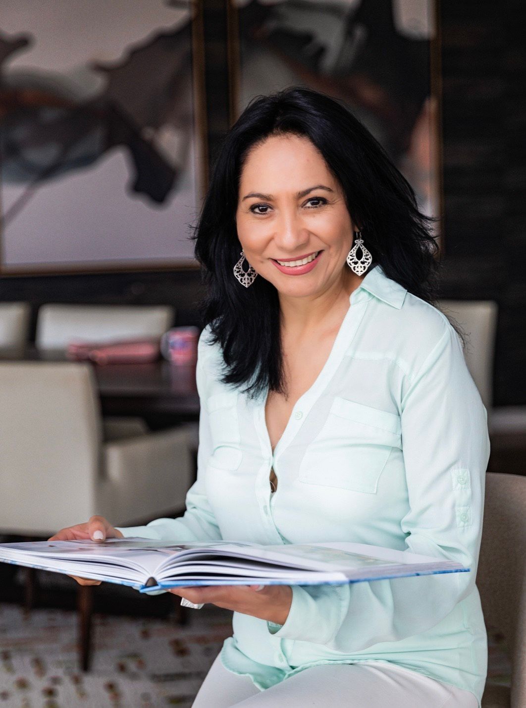 Woman smiling, holding open book, wearing light blue shirt, in a room with a painting.