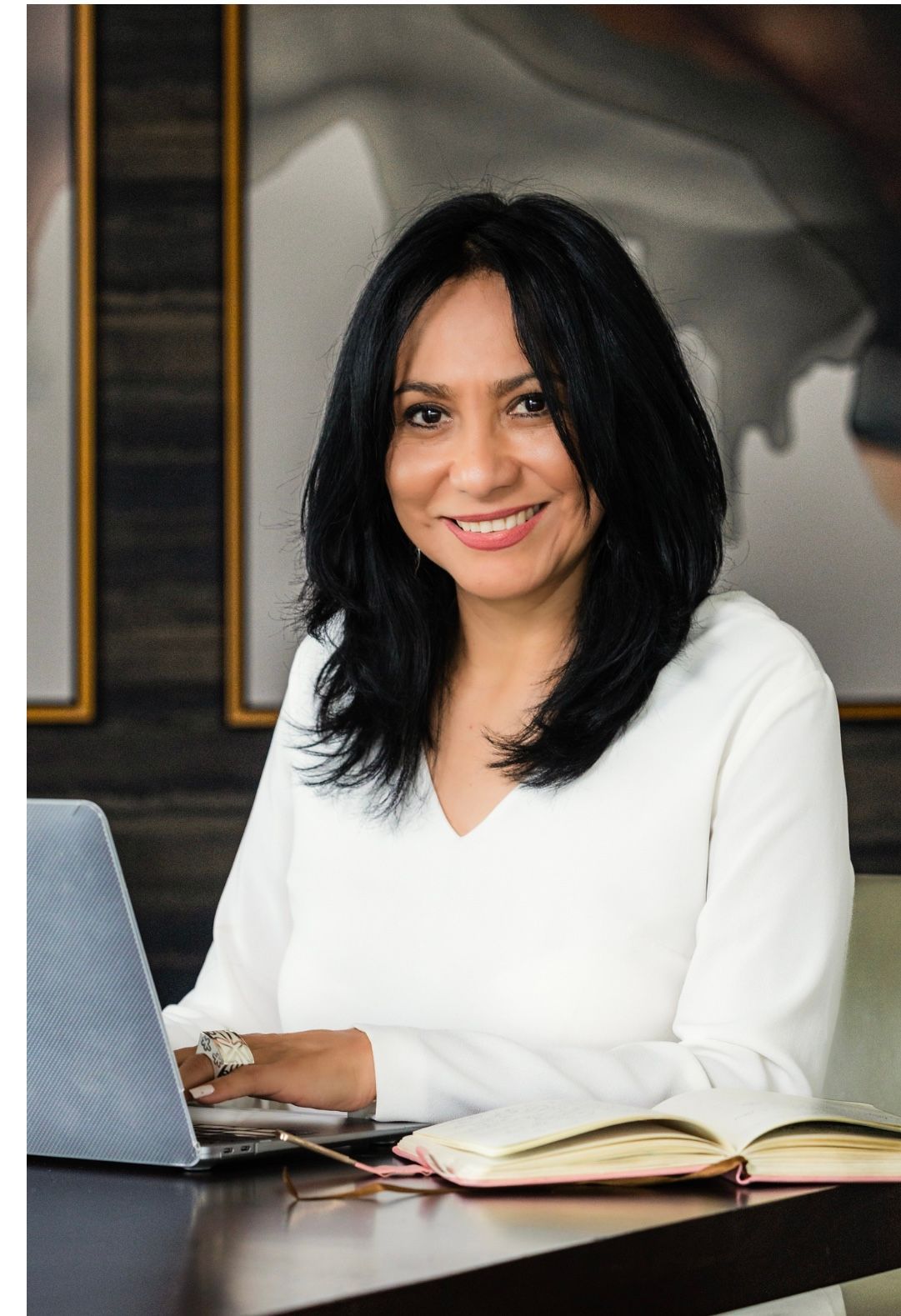 Woman smiling, working on laptop at desk with a notebook, wearing a white long-sleeved shirt.