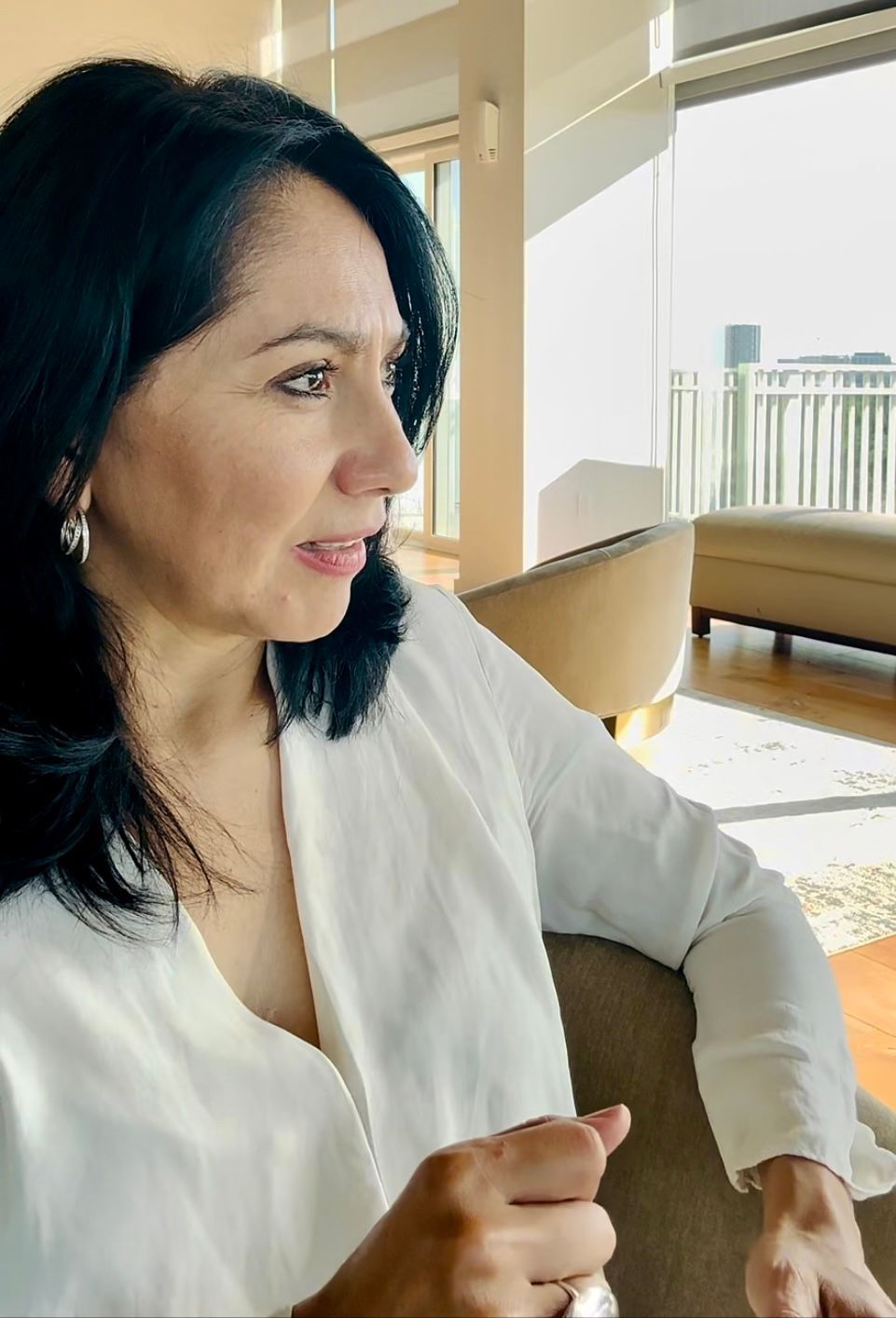 A person with dark hair and a white top sits in a brightly lit room, looking away from the camera.