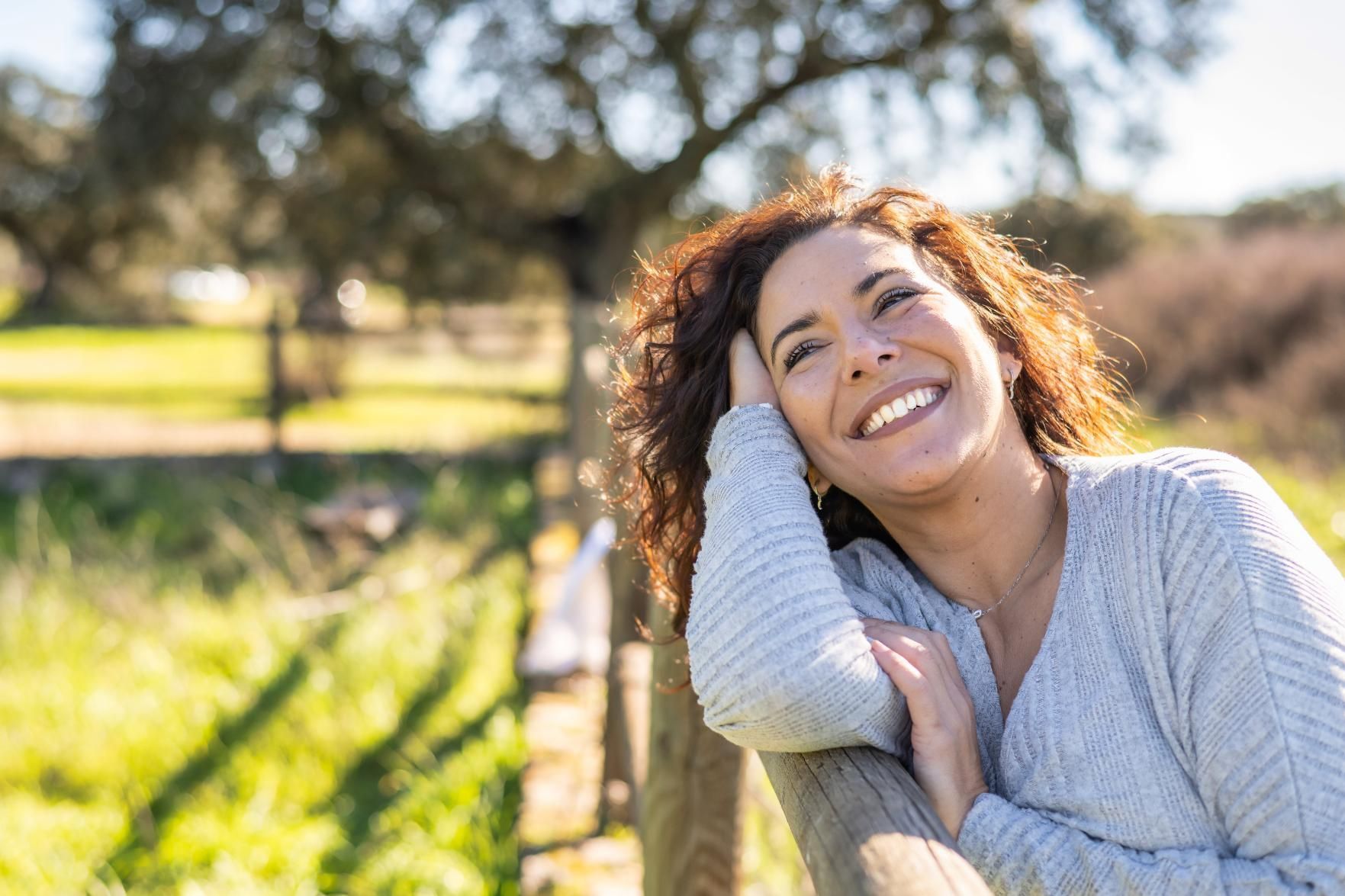 Woman leaning on fence, smiling and looking up outdoors, sunny day.