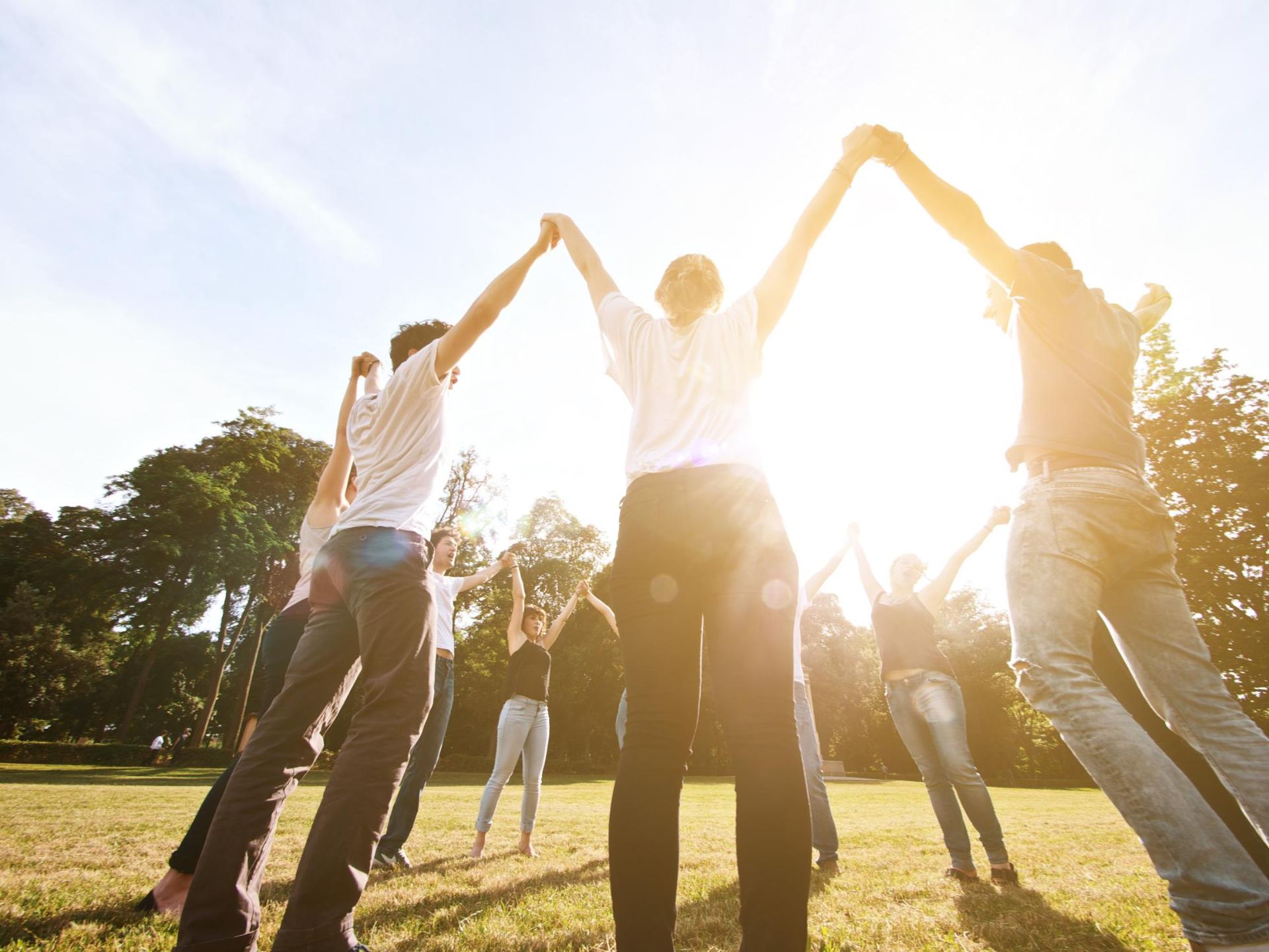 People standing in a circle with arms raised, holding hands against the sun in a grassy field.