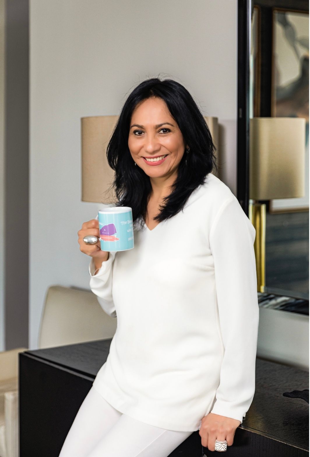 Woman in white outfit sitting indoors, smiling and holding a blue mug.