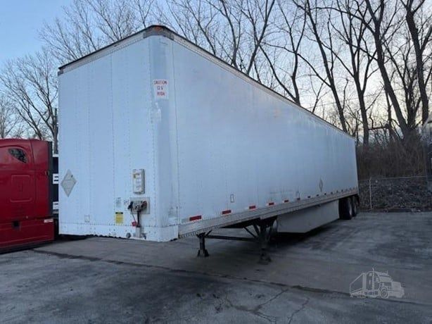 White semi-trailer parked outdoors near leafless trees. A red truck cab is partially visible on the left.