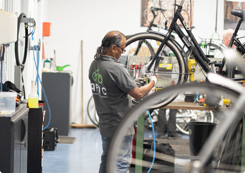 Man in gray shirt repairing a bicycle wheel inside a bike shop.