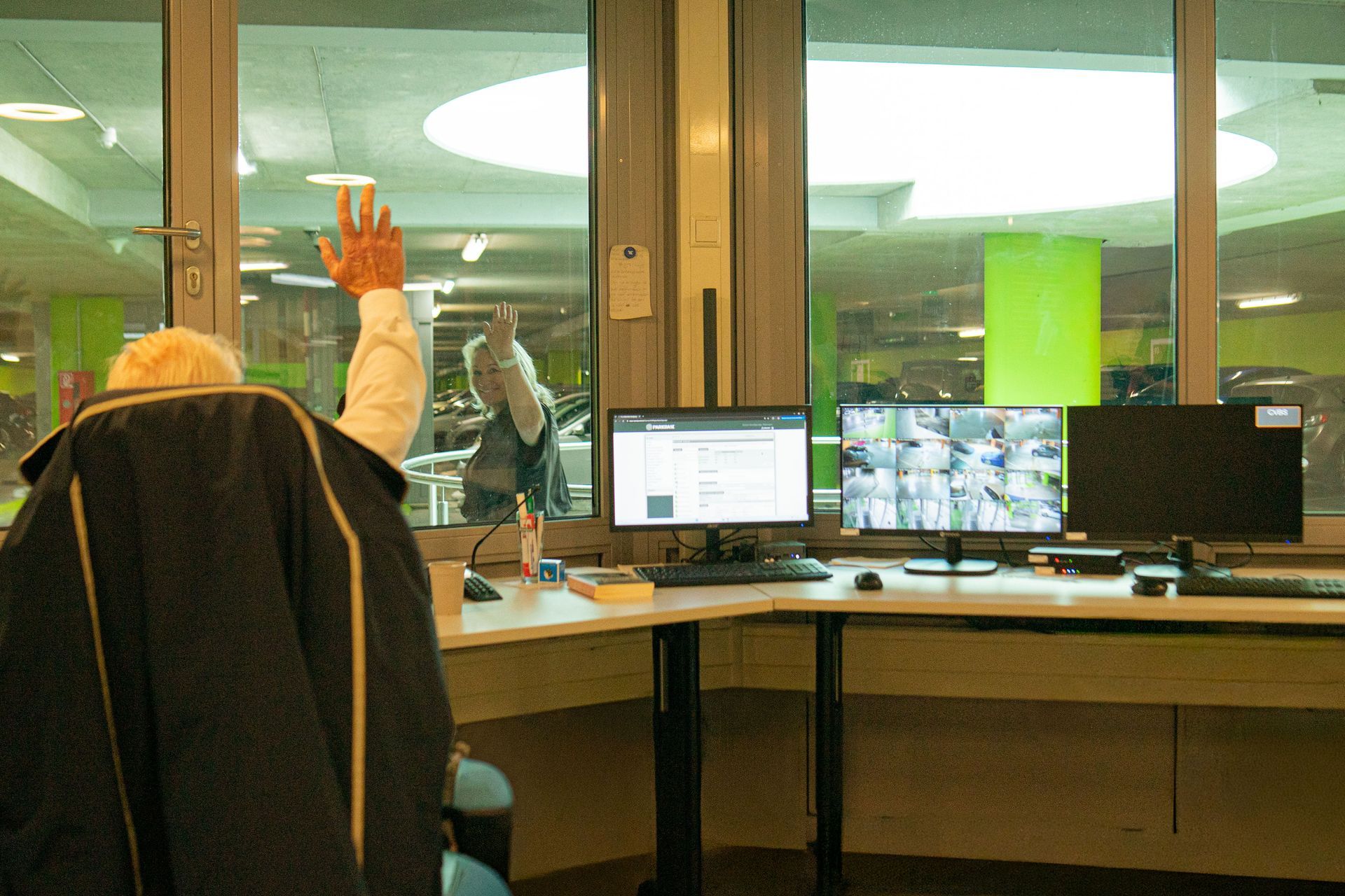 Een parkeerbeheerder  in zijn kantoor achter beeldschermen in een parkeergarage. De parkeerbeheerder zwaait naar een passant