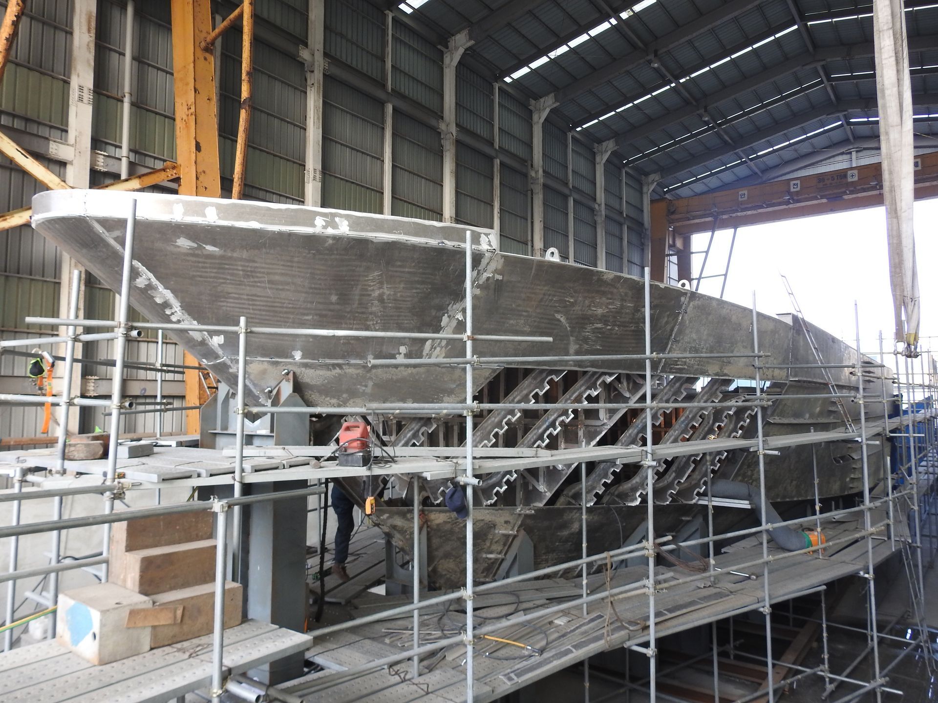 A large ship under construction in a metal workshop, with scaffolding, workers, and tools.