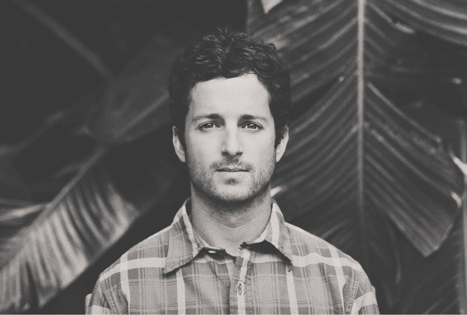 Man with curly hair in plaid shirt, neutral expression, in front of tropical plant leaves, black and white.