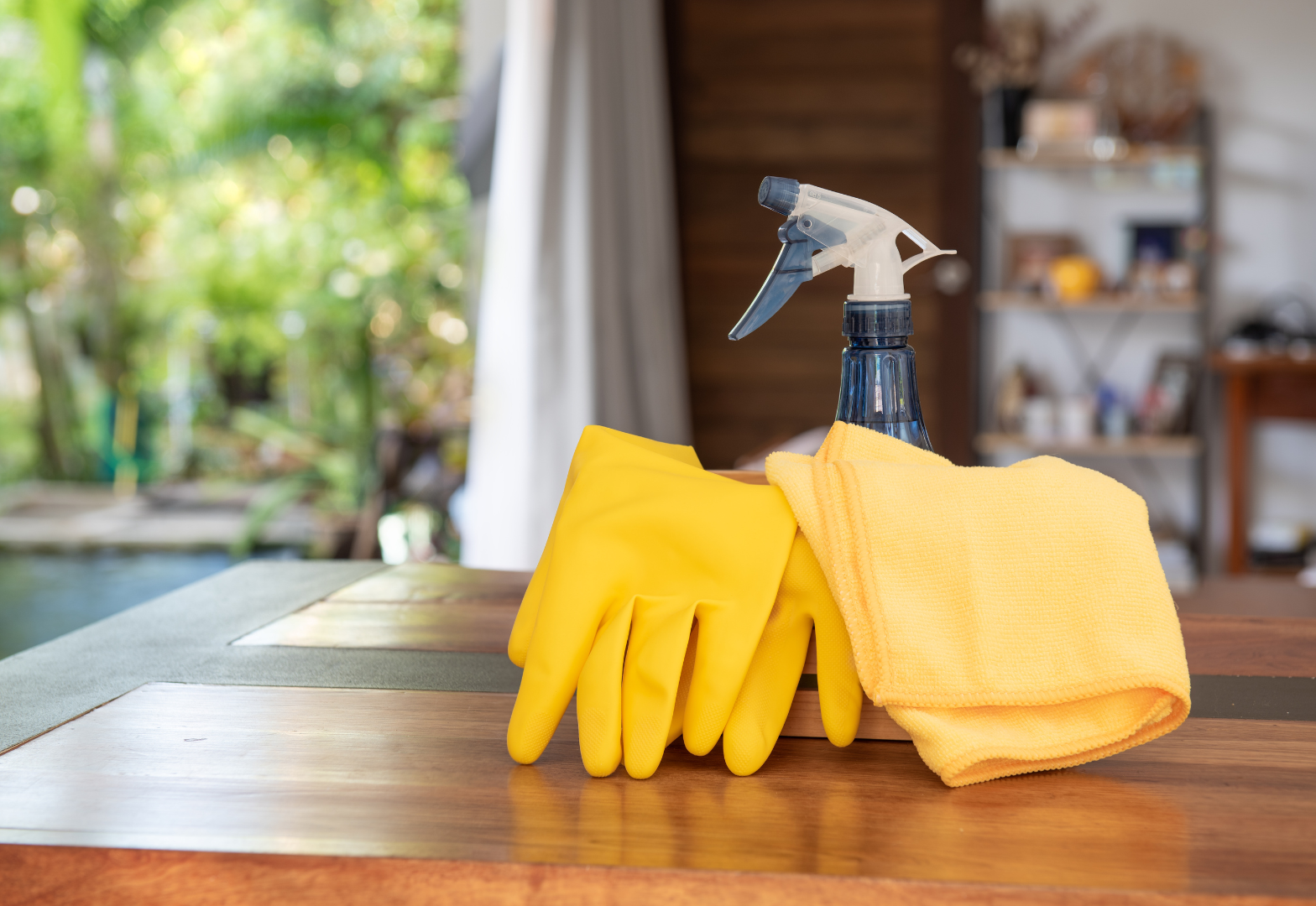 Yellow cleaning gloves and cloth beside a spray bottle on a wooden table near a window