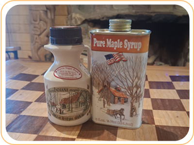 Two syrup containers: ceramic jug and metal tin with winter scene label.