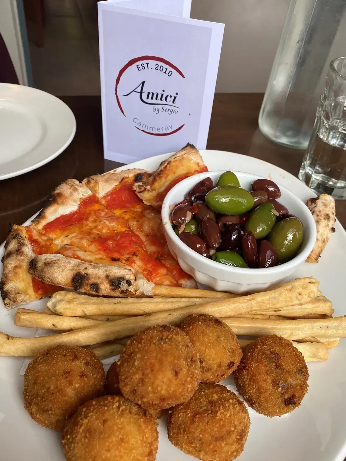 A plate of food with a pizza , olives , and bread sticks on a table.