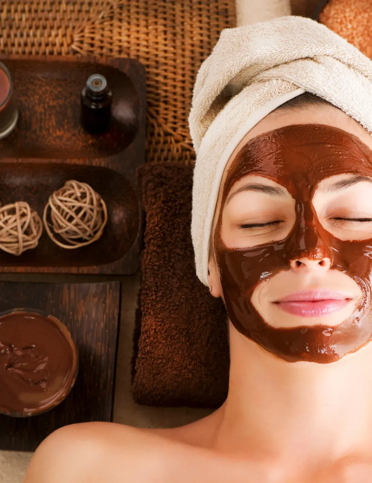 A woman with a towel wrapped around her head has a chocolate mask on her face.