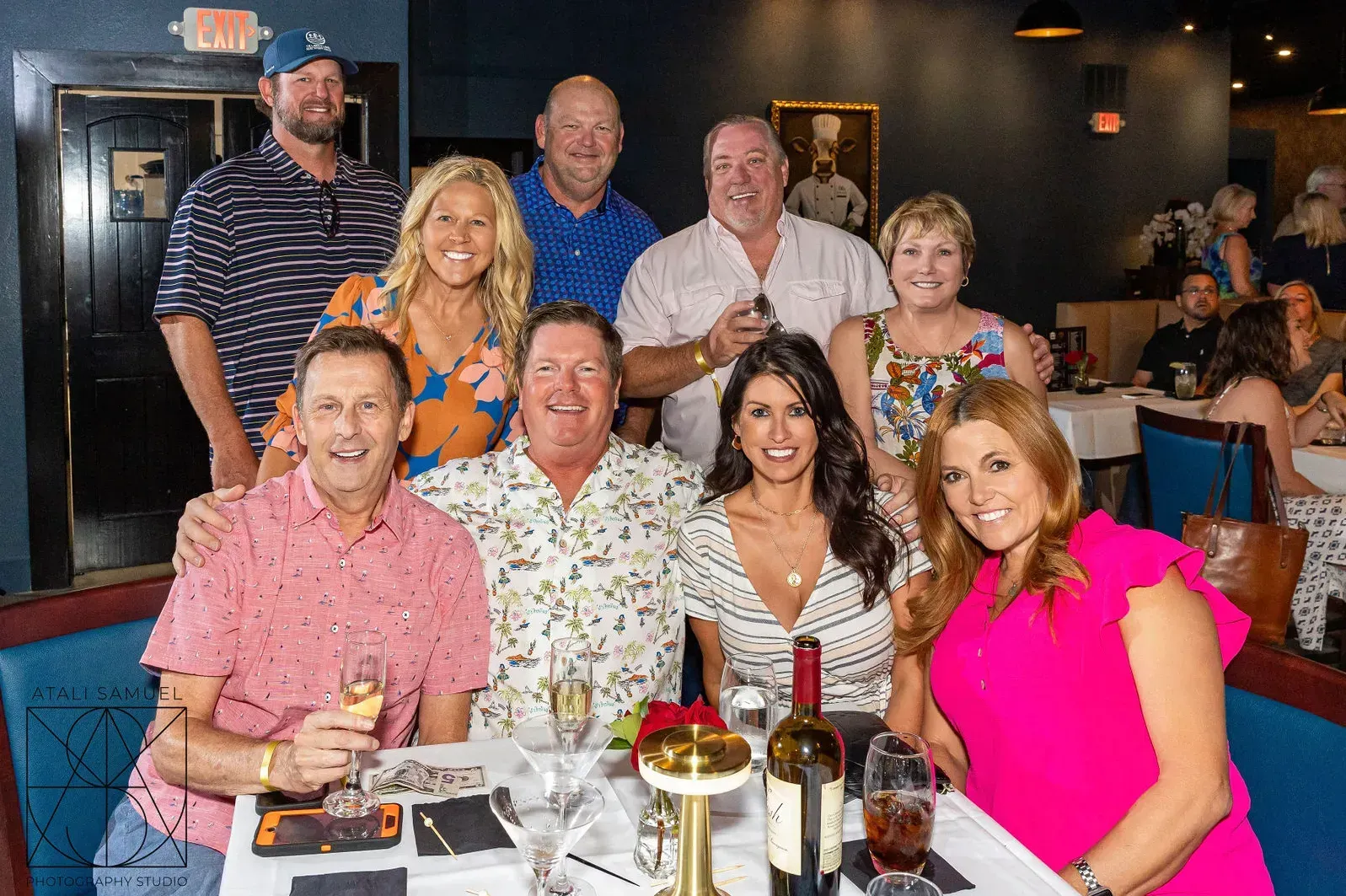 Group of people smiling at a table in a restaurant; glasses and wine bottles present.