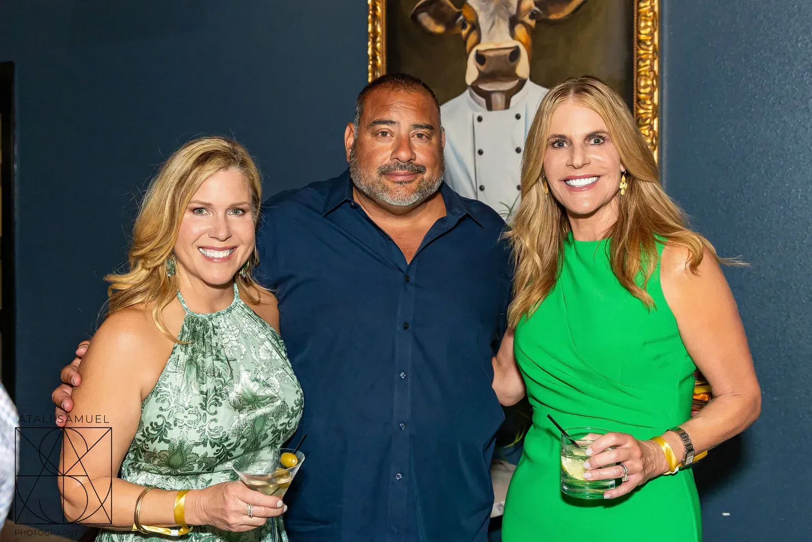 Three people smiling, posing together indoors; man in blue shirt between two women.
