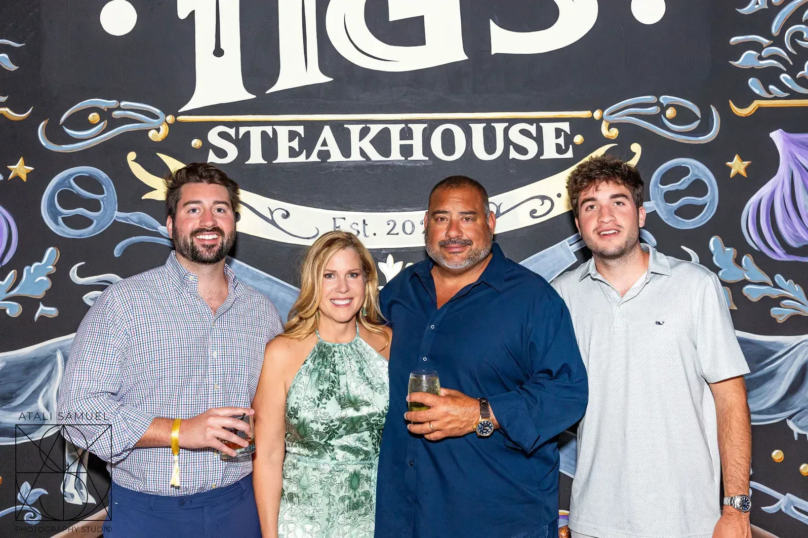 Four people pose in front of a steakhouse sign. Two hold drinks; all smile. Restaurant background.