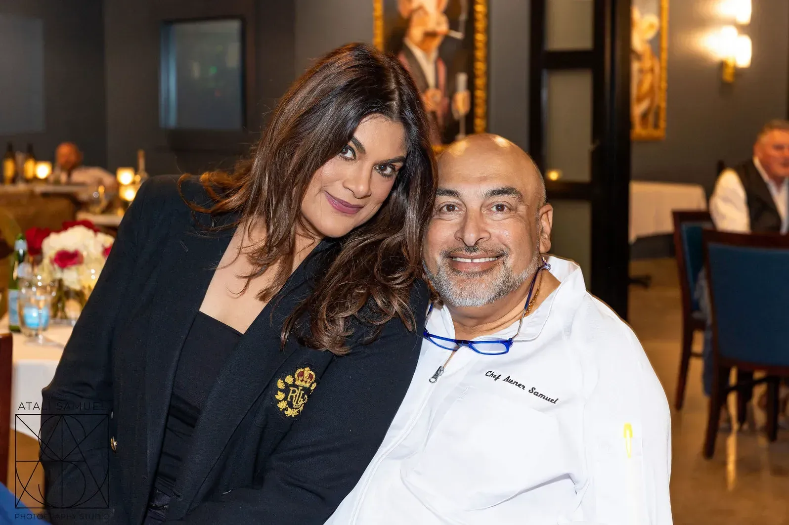 Woman in black blazer smiles with man in white chef's coat, seated in restaurant.