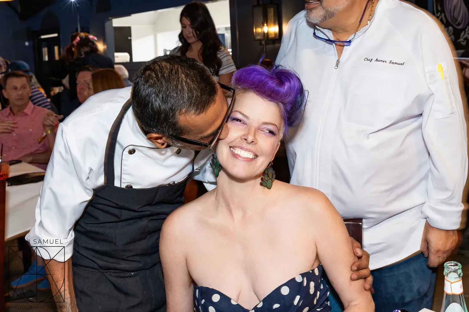 Chef kissing a woman with purple hair at a restaurant table; two men smile nearby.