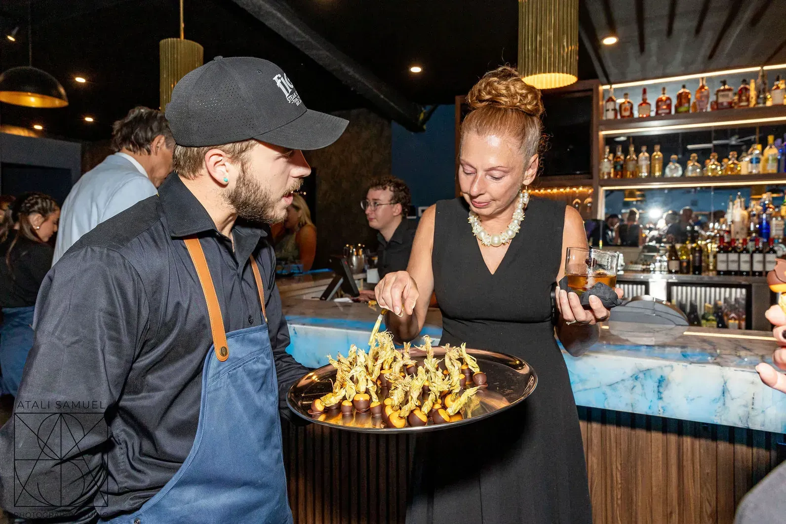 A server and woman with a tray of food near a bar. The server wears an apron and cap.