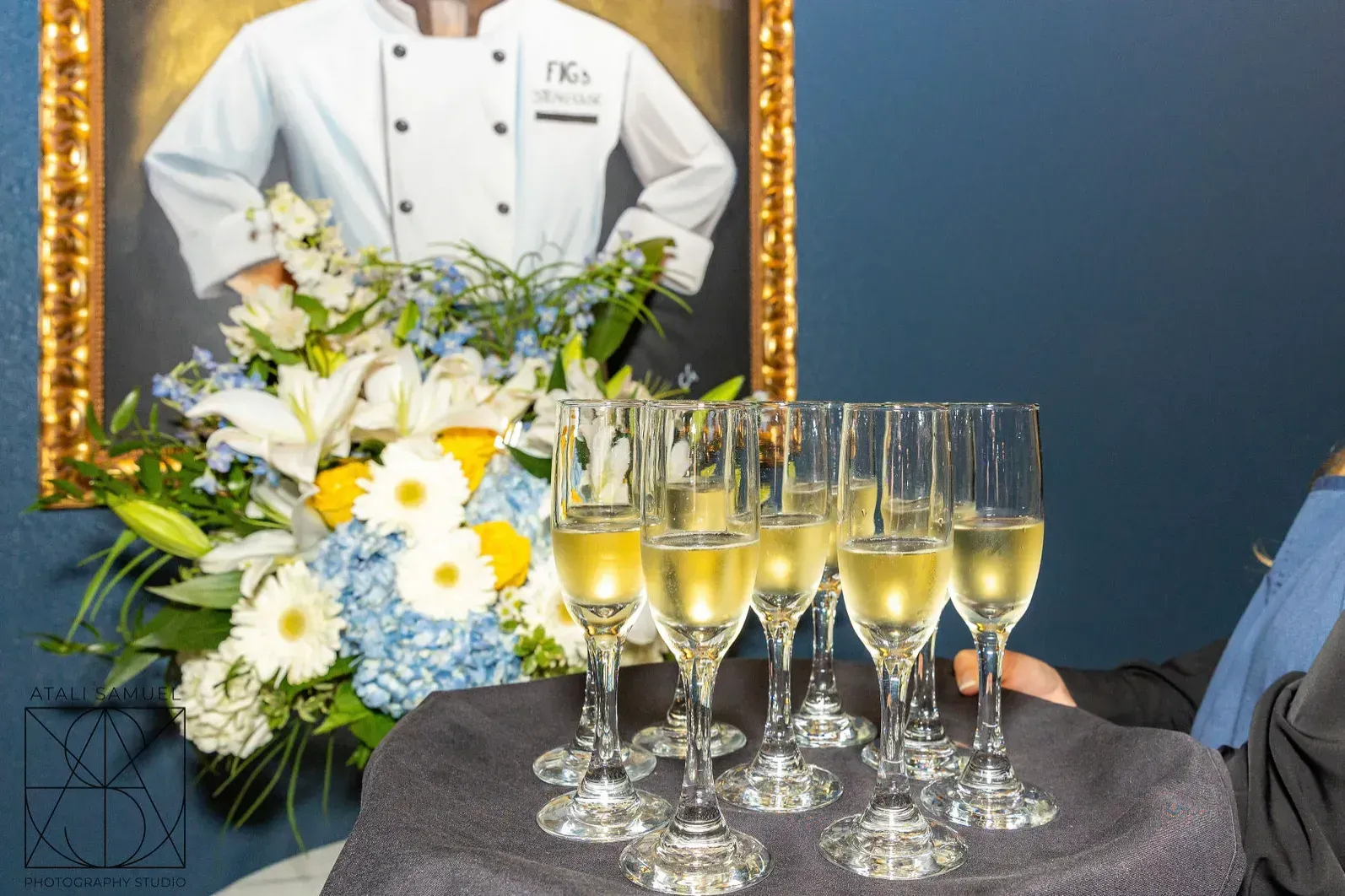 Champagne flutes on a tray, next to a floral arrangement and a portrait of a chef.