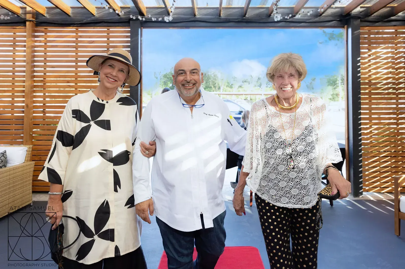 Three people smiling, one man with arms around two women, walking outdoors under a pergola.