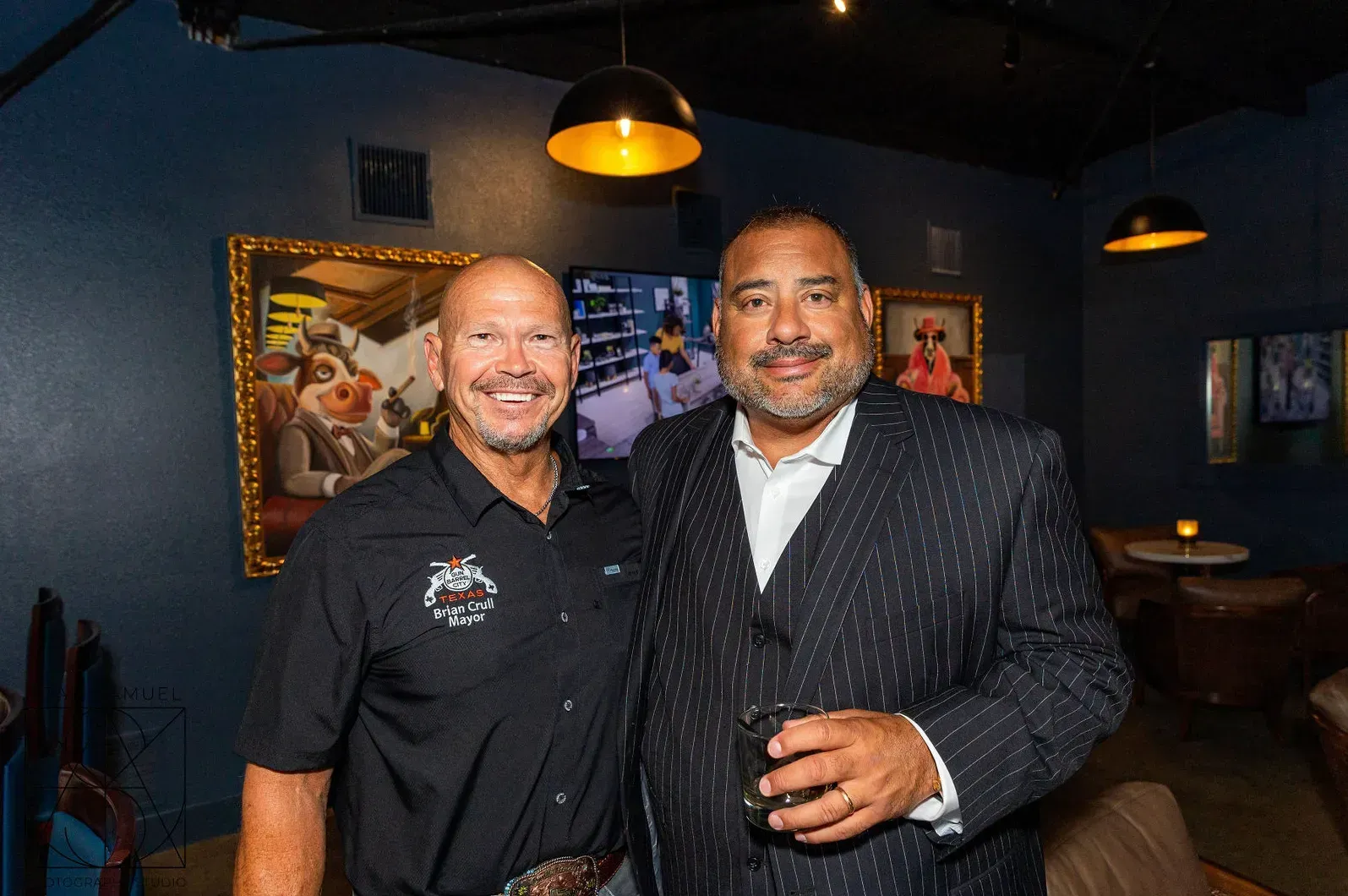 Two men smiling, posing together inside a dimly lit bar. One in a black shirt, the other in a suit.