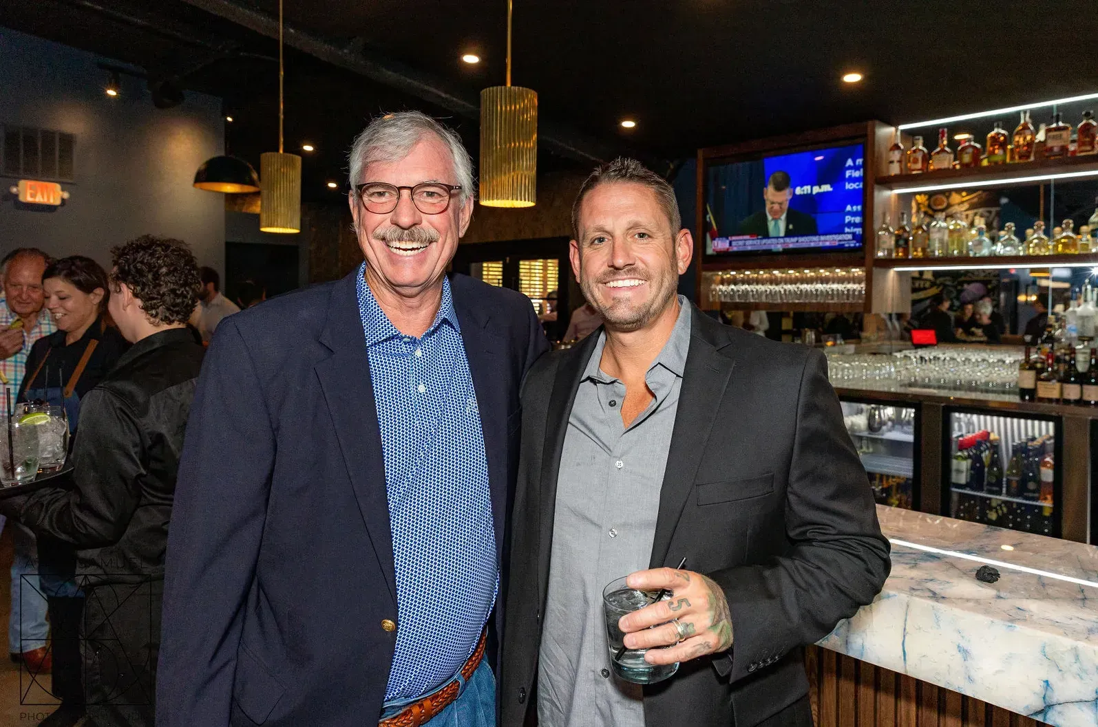 Two men smiling, arm-in-arm, in a bar. One wears a blue blazer, the other a black jacket.