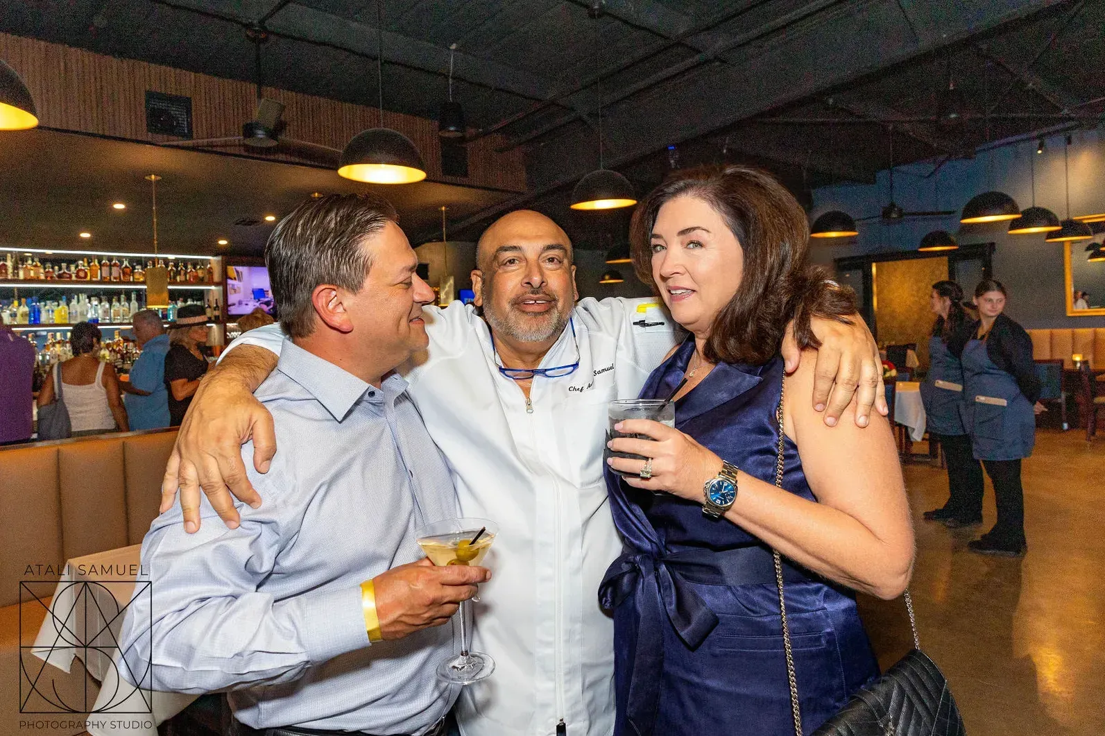 Three people embracing at a restaurant: one man in a blue dress shirt and another in a white jacket, woman in a blue dress.