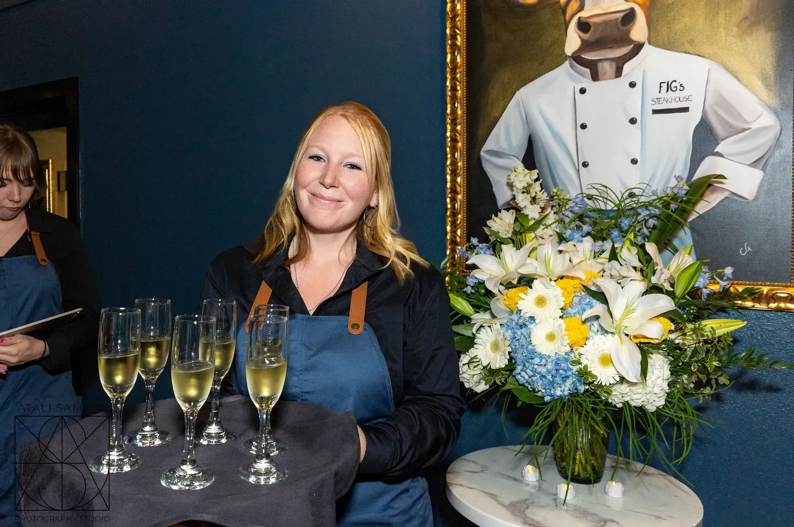 Woman holding a tray of champagne glasses smiles, with flowers and a chef-cow painting in the background.