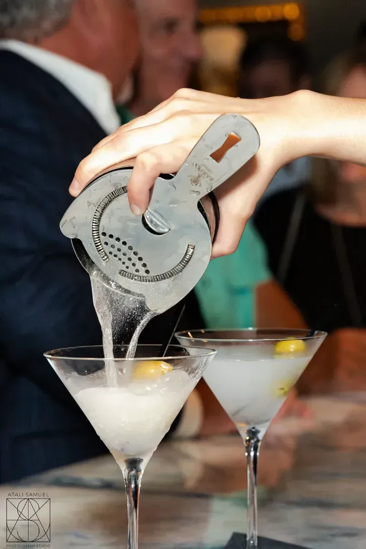 Bartender pouring a drink from a strainer into two martini glasses.