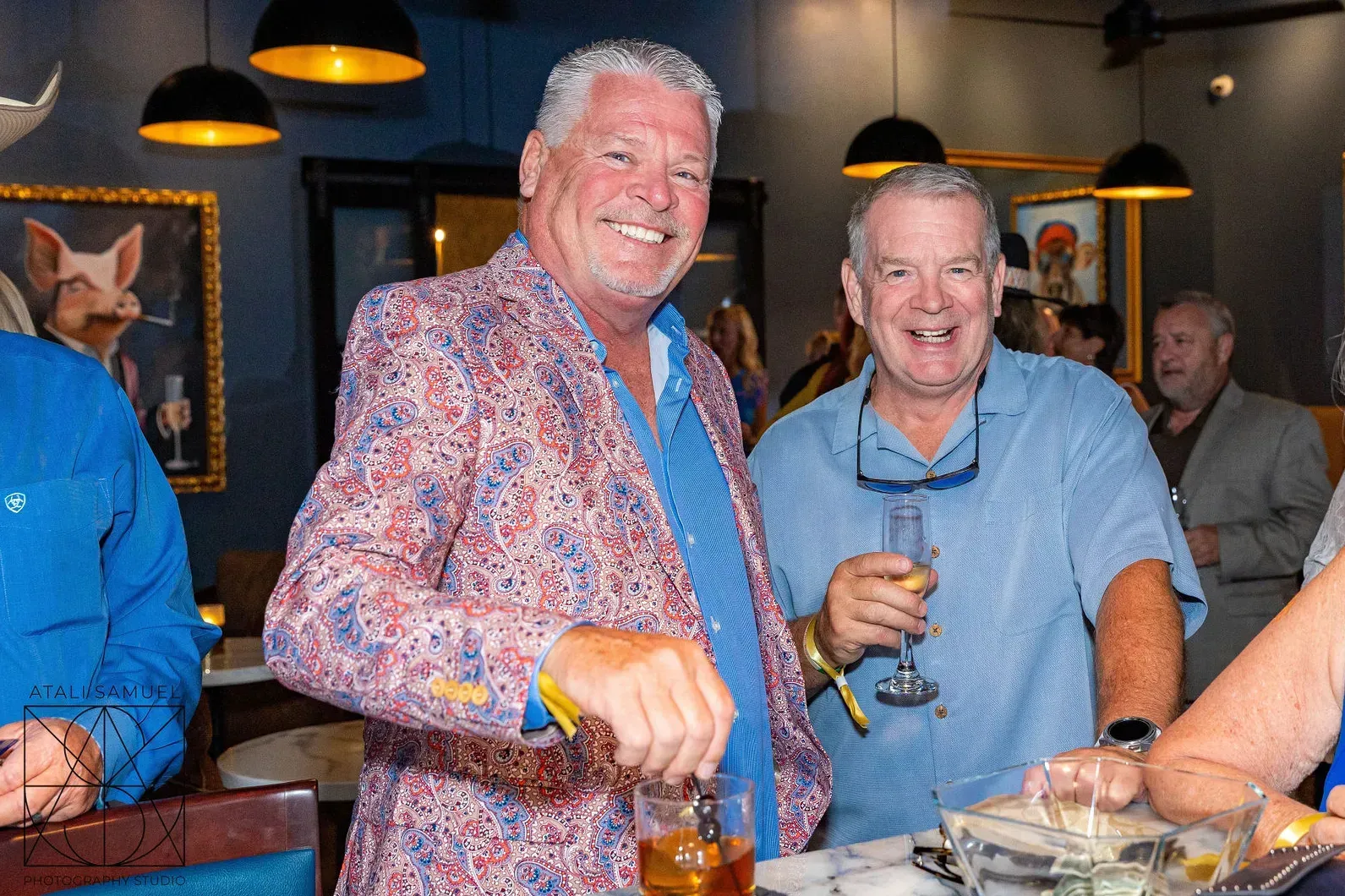 Two men at a bar, one in a colorful jacket smiling, the other holding a drink, smiling.
