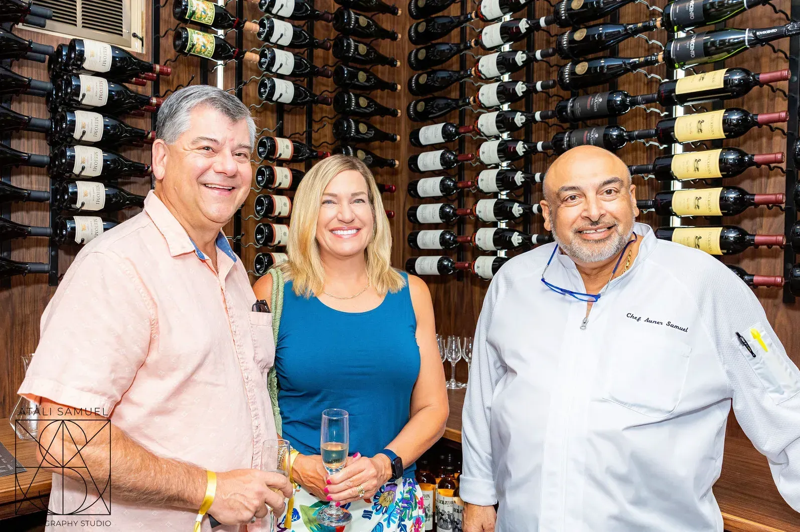 Three people smiling in front of a wine rack. One in chef's whites, the others in casual wear, holding drinks.