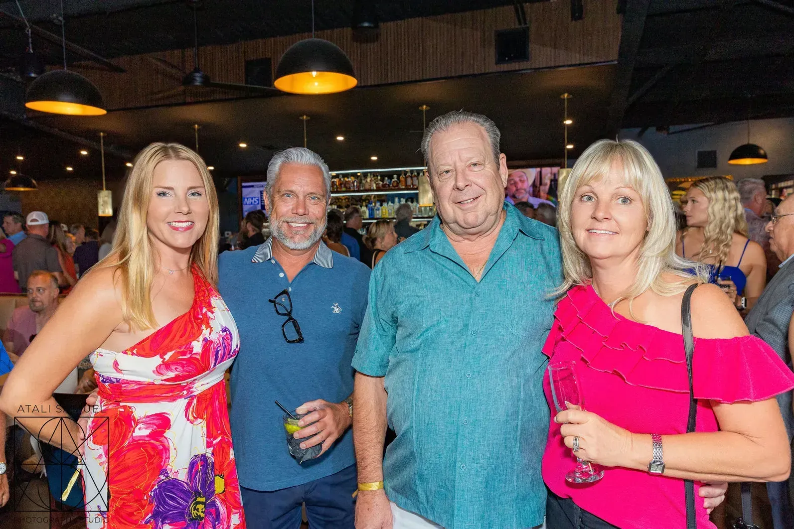 Four people smiling at an event; a woman in a red dress, three others with drinks.