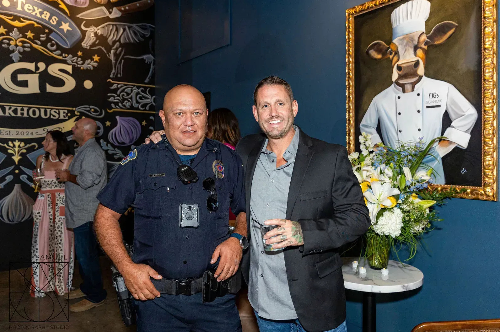 Two men pose inside a restaurant: one in a police uniform, the other in a suit, with a painting of a cow in a chef's outfit nearby.