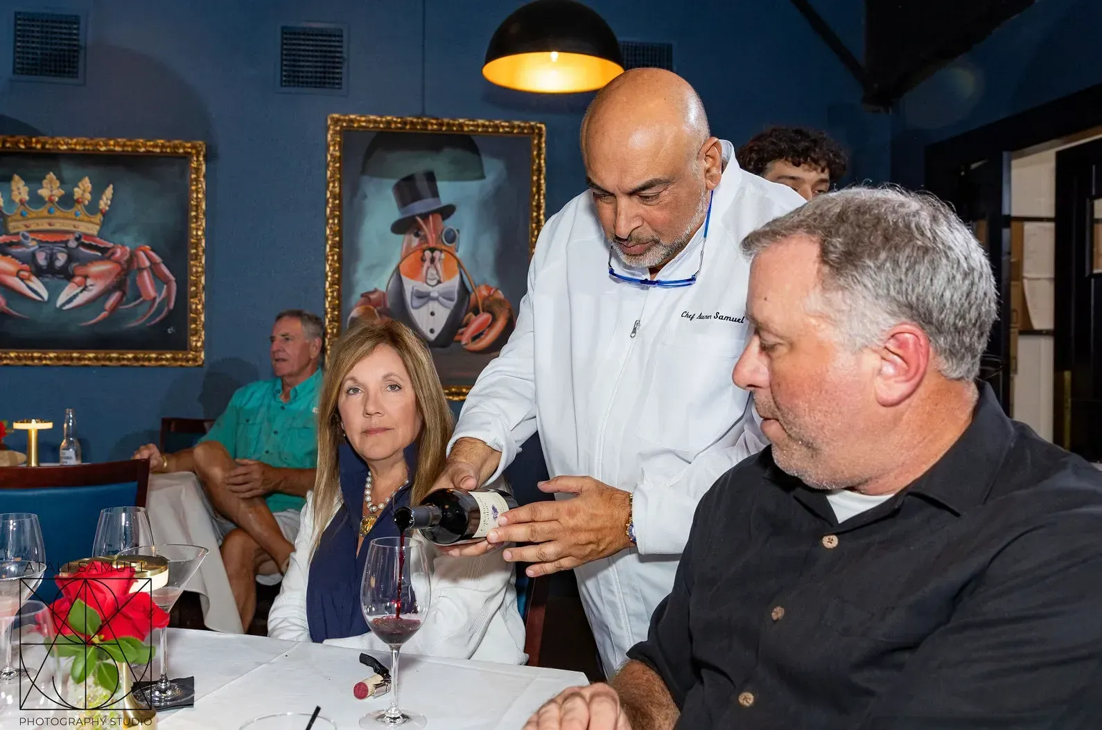 Chef pours wine for a couple at a table; blue walls, paintings of crabs in a restaurant setting.