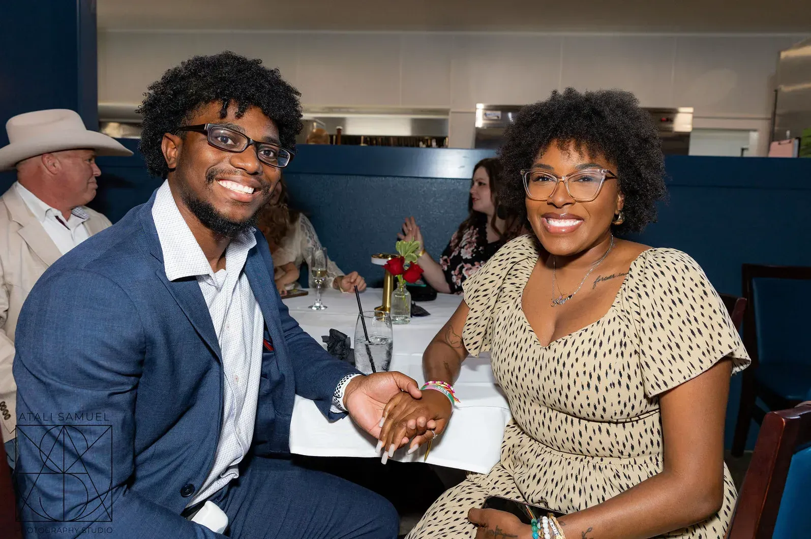 Smiling couple holding hands at a restaurant table. They are in semi-formal attire, with other diners in the background.