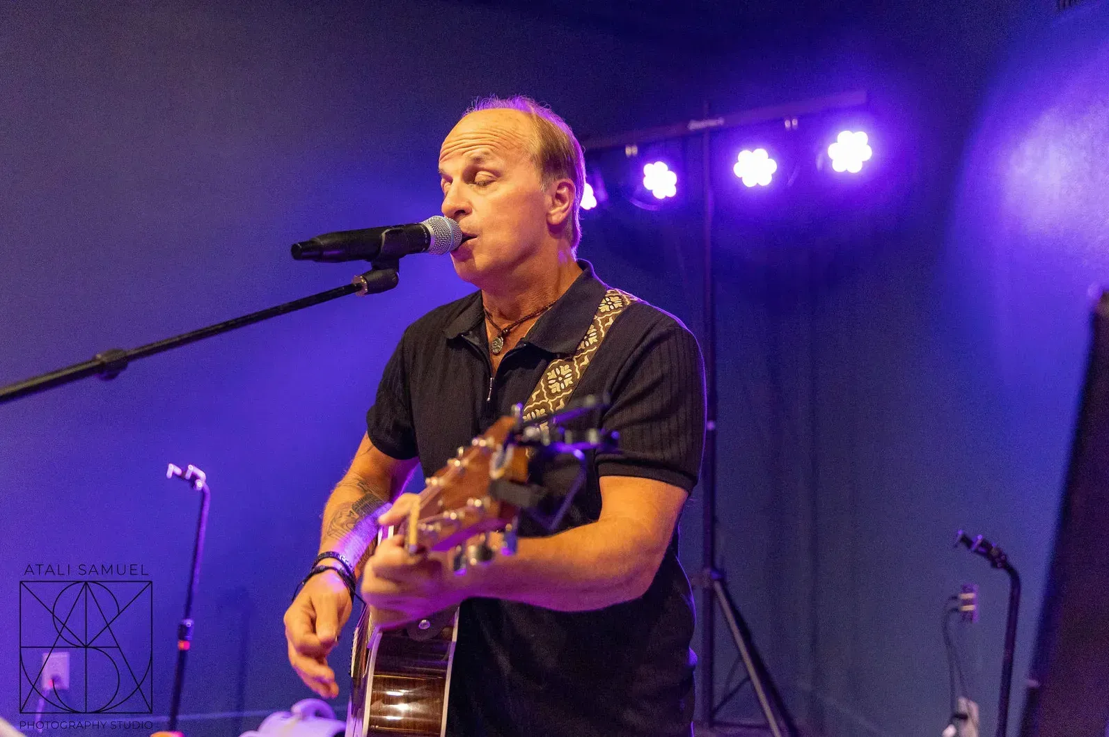 A man singing and playing acoustic guitar on a stage with a microphone. Purple lighting.