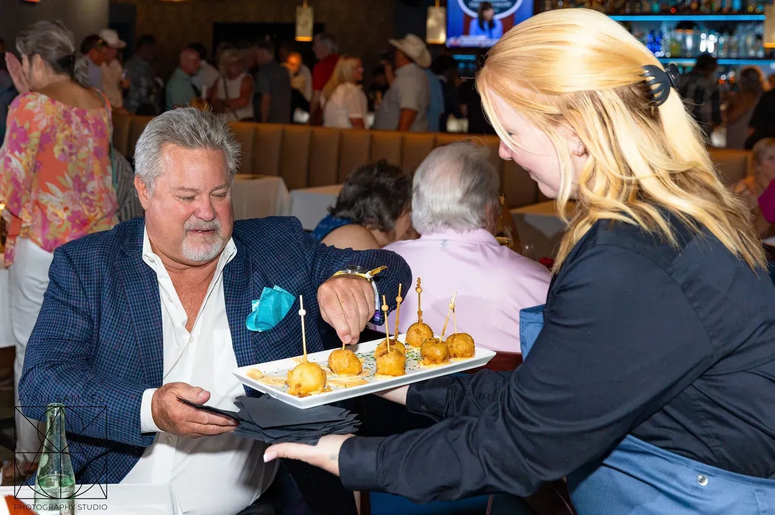 Man in blue blazer taking appetizer from a server in a restaurant. Others dine in background.