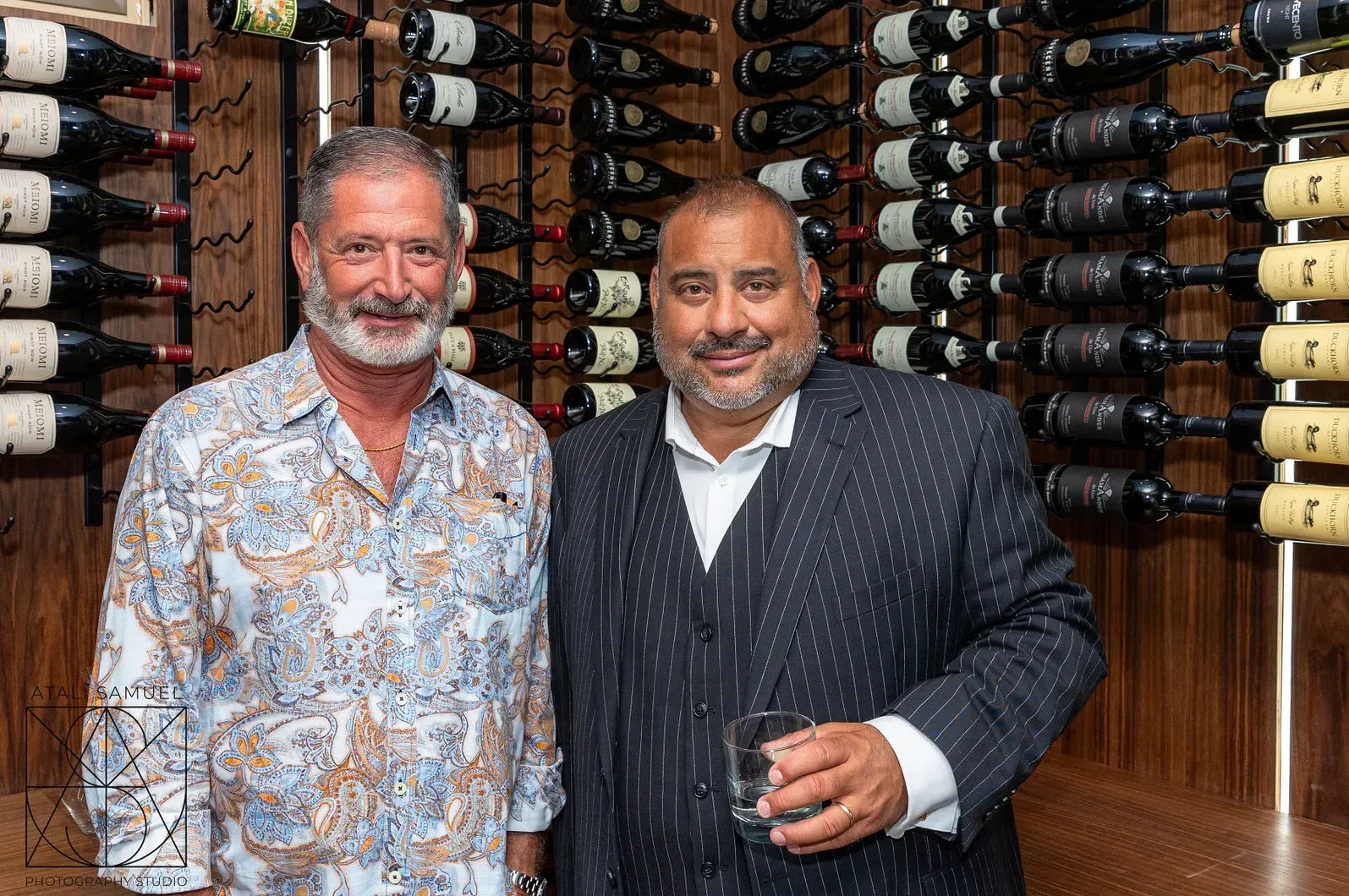 Two men stand in front of a wine rack. One in a patterned shirt, the other in a suit, holding a glass.