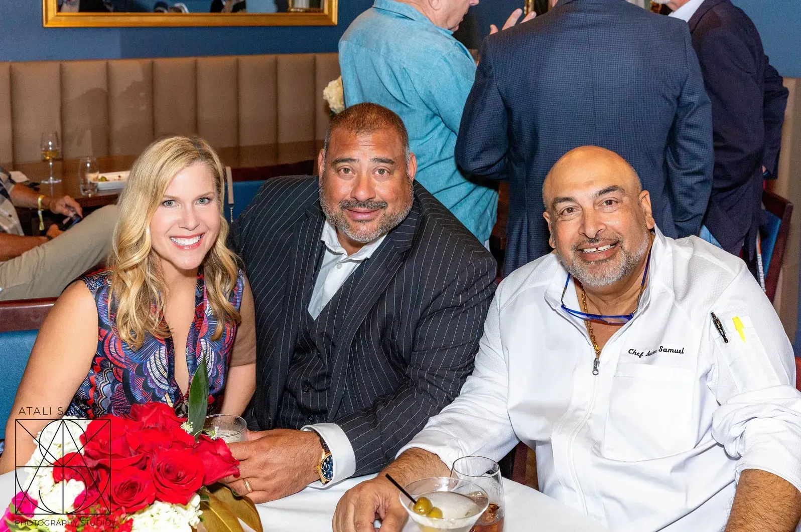 Woman with two men at table, red roses, man in chef's coat, other guests in background.