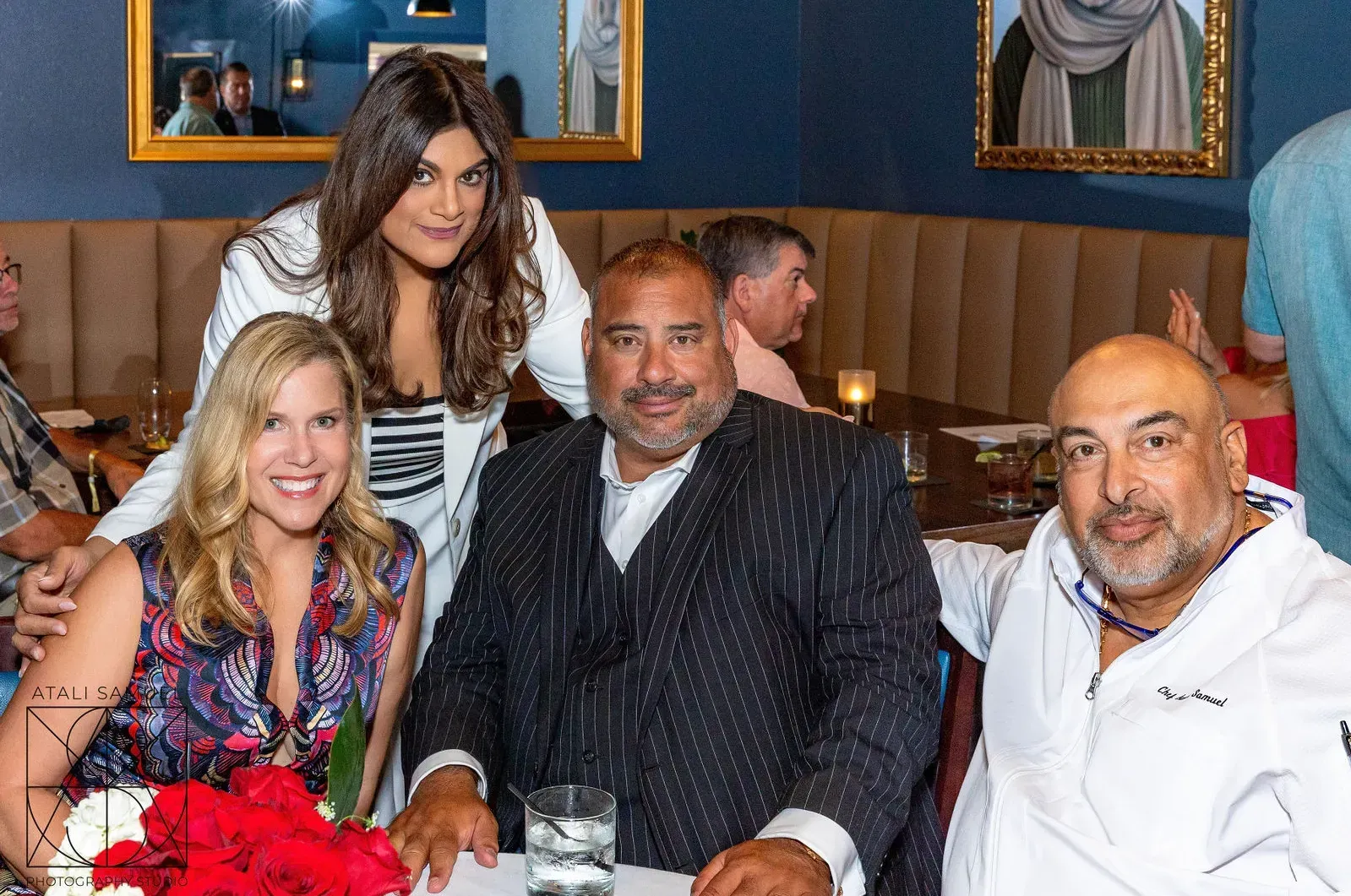 Four people at a table in a restaurant. A woman stands behind the others. They appear to be smiling and enjoying a meal.