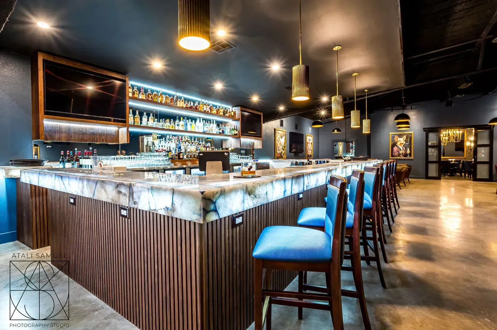 Upscale bar interior with marble bar, blue chairs, and liquor bottles on shelves.